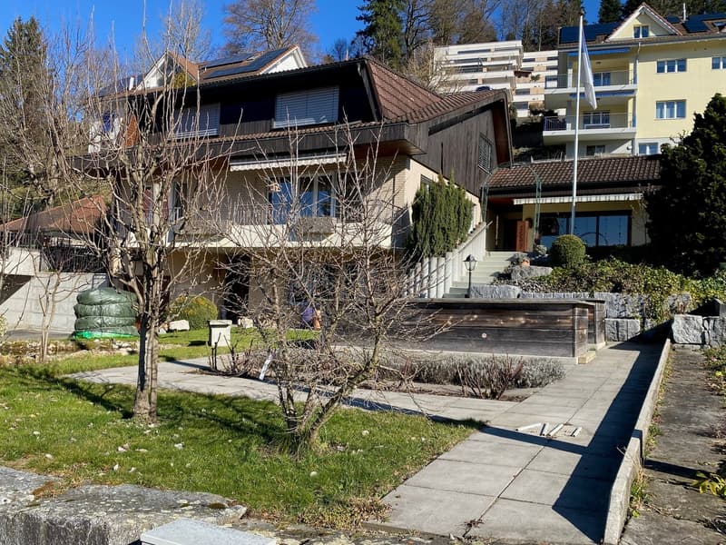 House with gable roof, surrounded by trees, large front garden, raised ground floor, view of the hills
