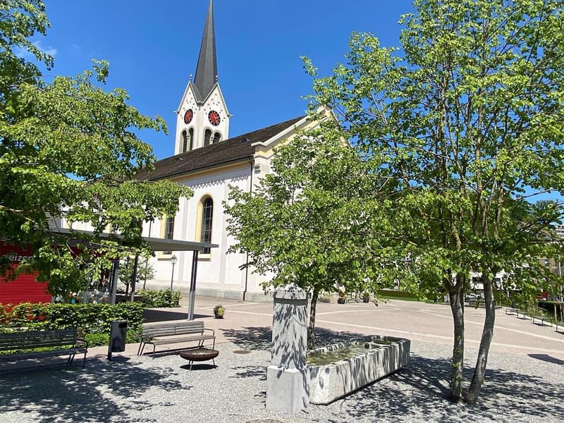 church with spire, red clock faces, trees, benches, small fountain