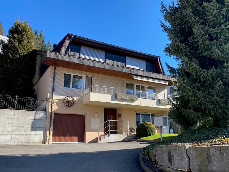 Two story house, beige facade, large balcony, brown garage, black metal railings, green trees