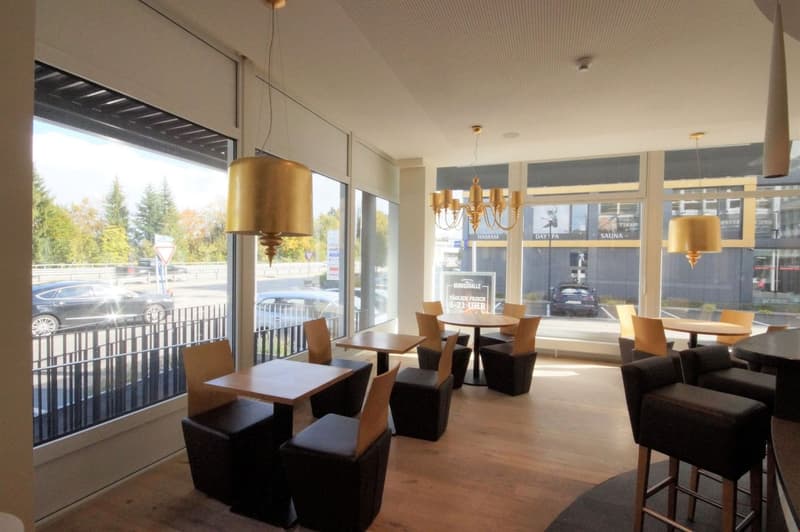 Spacious dining area with large windows overlooking a parking lot, wooden tables and chairs, black leather stools, gold pendant lights, and signage for a sauna and other amenities.