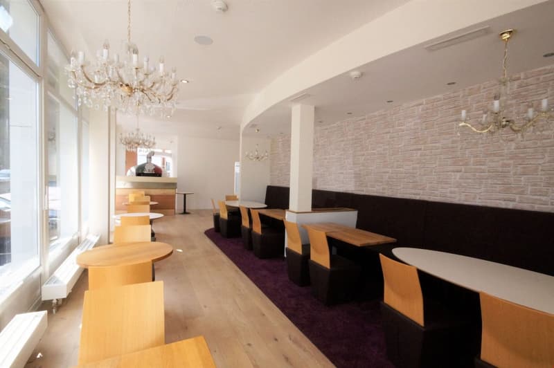 The image shows a spacious dining area with a brick wall, wooden tables and chairs, and a large chandelier. There is a fireplace visible in the background. The overall decor appears to be modern and elegant, creating a cozy and inviting atmosphere.