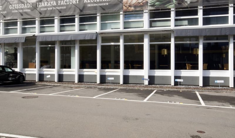 A commercial building with large glass windows, a parking lot in front, and signage for the Izakaya Factory restaurant and Geissdadi Schwizer Chucht.