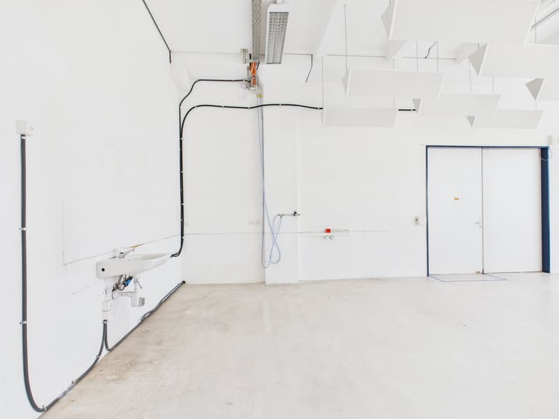 The room is completely empty, with no visible features or items. The walls are white, and the floor appears to be made of a white material. On one wall, there is a mounted sink, with a pipe connected to it. There are also wires hanging from the ceiling, p