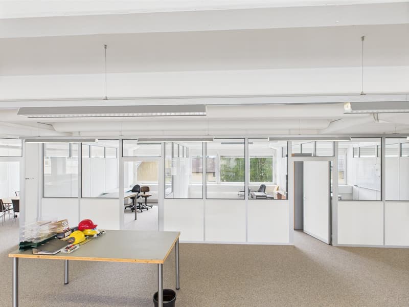 Open space office with white walls, glass partitions, carpeted floor, fluorescent ceiling lights, a table with various items, and a trash bin.
