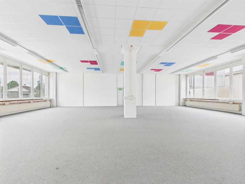Empty office space with white walls and gray carpet, large windows, and a central white column, color accents on the ceiling in blue, yellow, and pink.