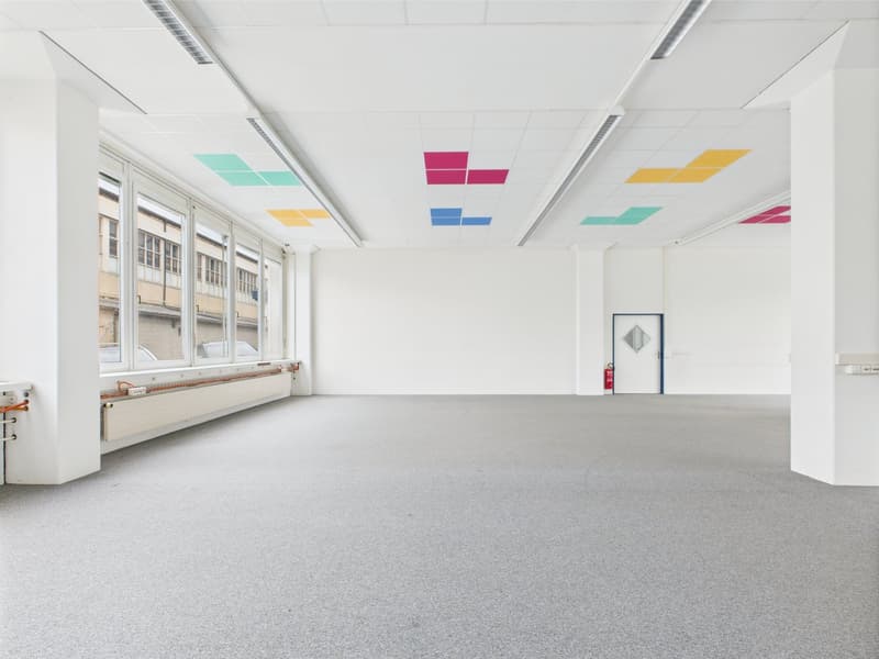 An empty room with white walls, ceiling, and floor, large windows, a white door with a white square on it, fire extinguisher nearby, a long white pillar, a white radiator, a ceiling with square lights.