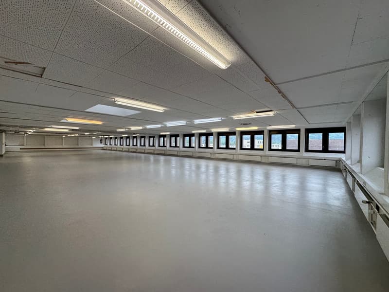 large open space with multiple windows along one wall, bright white ceiling with visible metal beams, fluorescent lighting, no visible furniture or decor