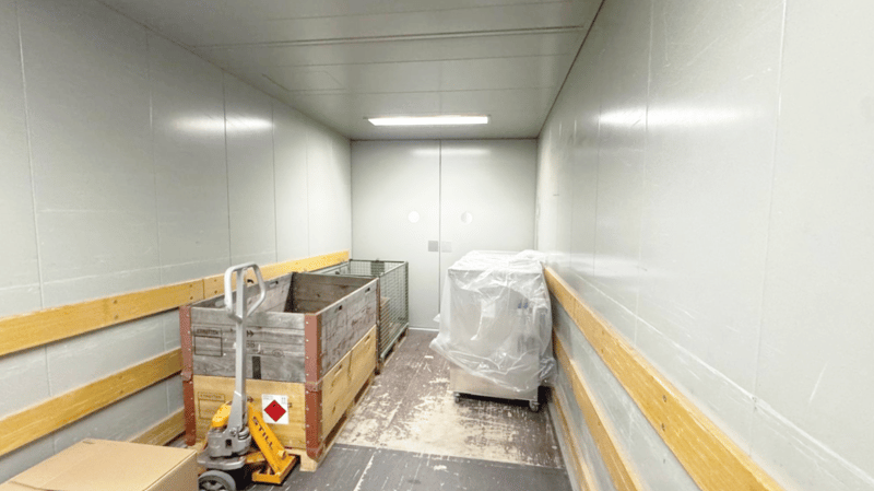 white walls, ceiling with recessed lighting, white floor, a covered appliance on a trolley, a wooden pallet covered in a tarp, a storage cage, a hand pallet truck