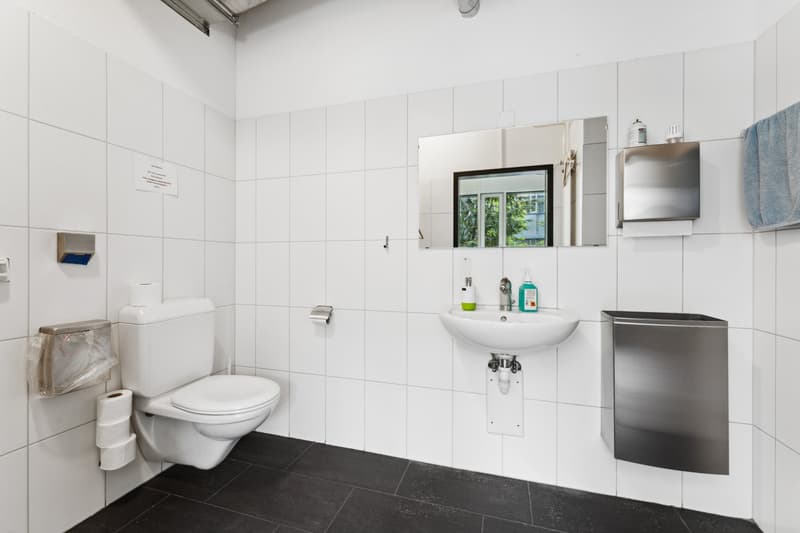 White tiled walls, black tiled floor, white toilet, sink, mirror, soap dispensers, towel holder with a blue towel