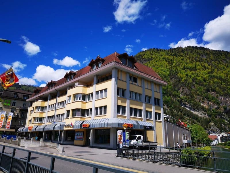 yellow building with red roof, multiple windows, located on a corner with mountains in the background