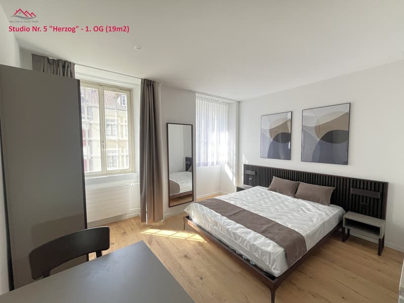 a room with a bed, a desk, a chair, a mirror, and art frames on the wall