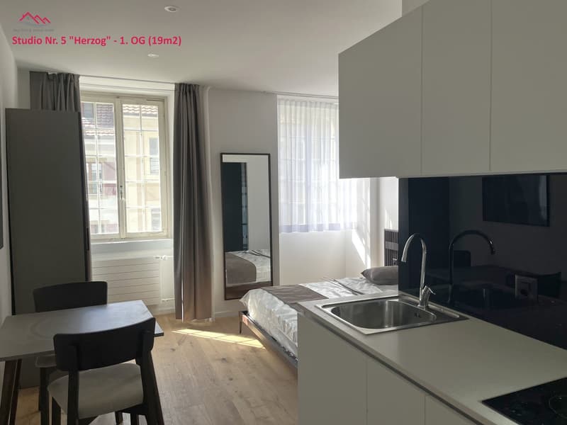 Small studio apartment with open kitchen, small dining table, bed, window, curtain, mirror, radiator, sink, faucet, stovetop.