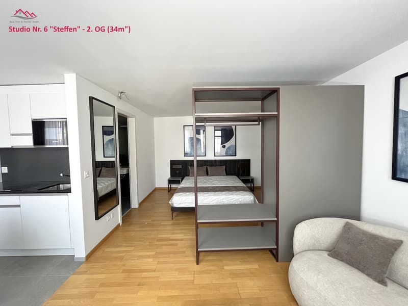 Compact room with wooden floor, bed, shelving unit, mirror, cabinet with sink, kitchen counter with stove, picture frames on the wall.