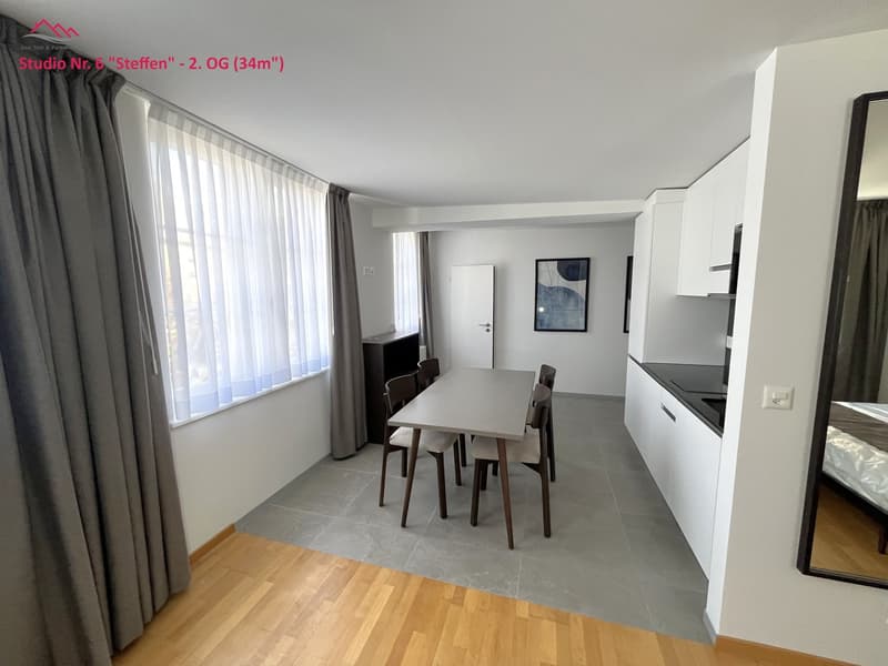 Dining area with table, chairs, open kitchen, wooden flooring, large window, mirror, bed, gray curtains, cabinets, power outlet