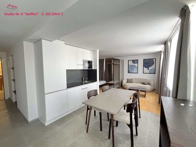 Open floor plan with kitchen, dining area, and living space; modern design; light grey flooring; white cabinetry; dining table and chairs; couch; bed; artwork on wall; wooden floor in living area