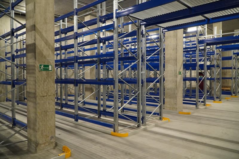 Steel storage racks, blue metal beams, yellow bumpers, concrete pillars, exit sign