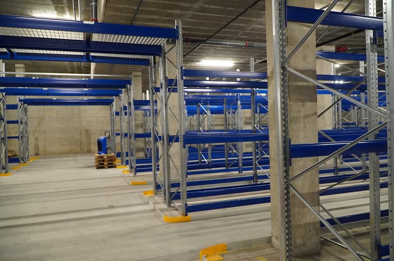 Warehouse with multiple levels of pallet racking, concrete floor, blue shelving, yellow edge protectors, storage box.