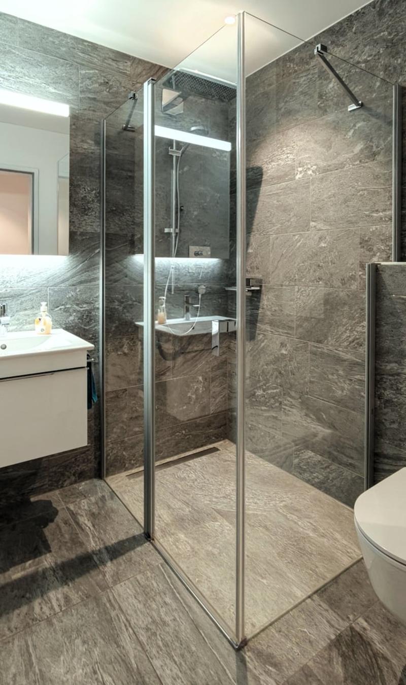 Gray tiled walls, modern glass shower stall, white countertop, white toilet, faucet, shower head, and shelves, mirror, sink