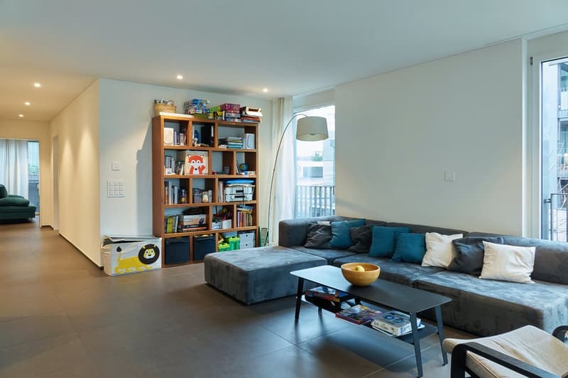 modern room with tile floor, a grey sofa, a coffee table with a bowl, a wooden shelf with books, a floor lamp and a balcony with a railing