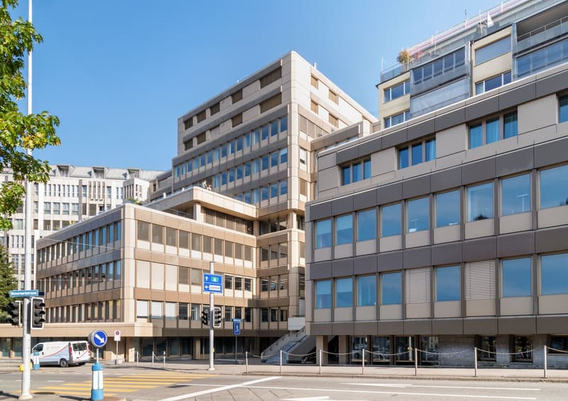 A large office building with multiple floors and many windows, some with white doors, situated on a raised ground floor