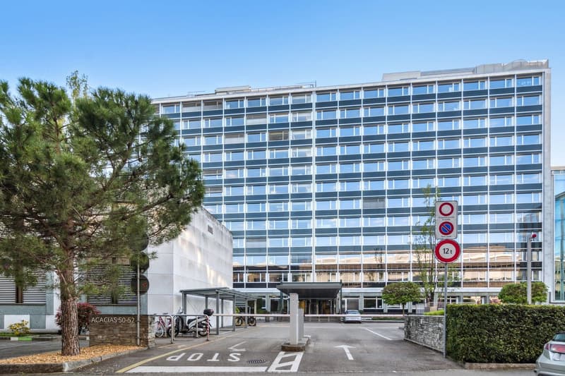 Modern office building, glass facade, multiple floors, entrance with canopy, parking area, bicycles parked, street signs, greenery