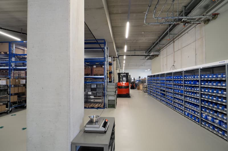 Warehouse with white flooring, multiple blue storage racks, forklifts, boxes, industrial weighing scale, metal pillars, and piped ceiling