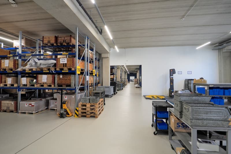 Warehouse with shelves filled with boxes and storage containers, a trolley for moving heavy items, white flooring, metal carts, bright lights on the ceiling