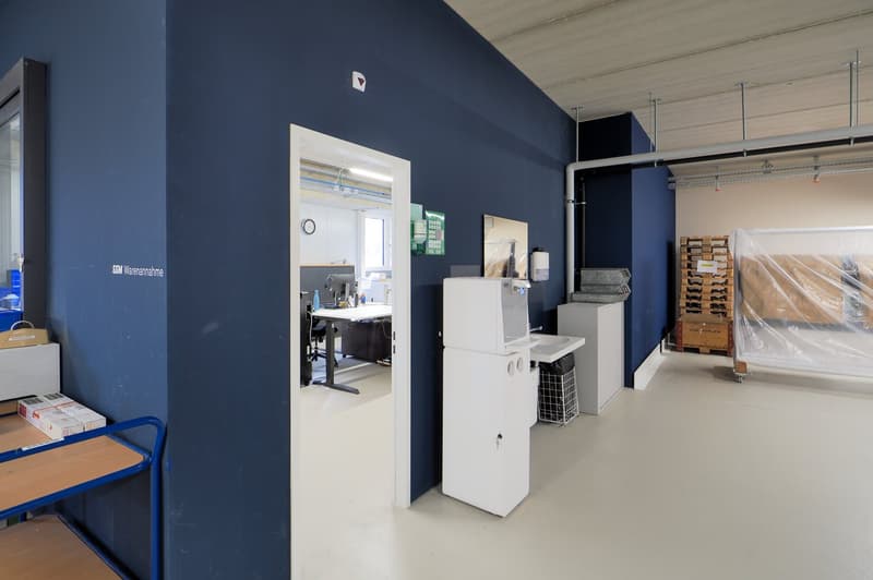 Open office space with a desk, monitor, chair, and clock. There is a storage box, a refrigerator, and a printer machine. There are also storage pallets and a plastic cover.