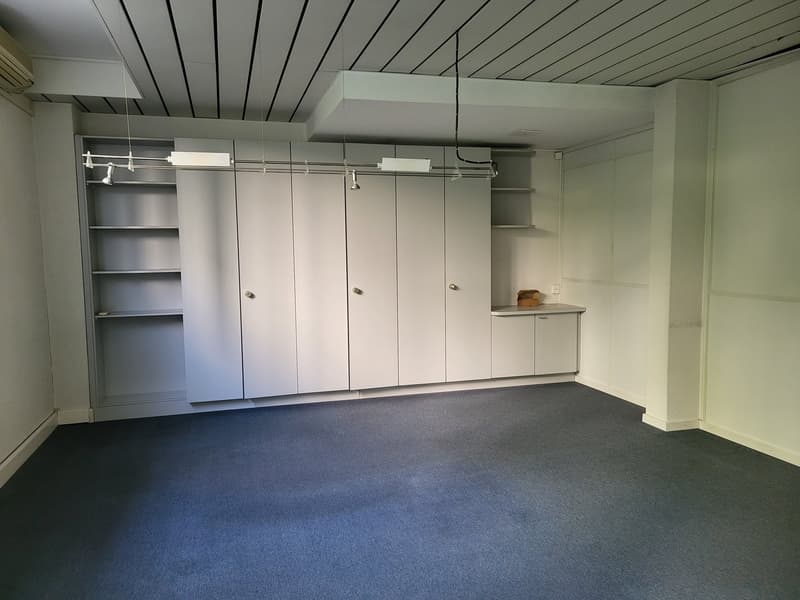 The room has a white ceiling, grey floor, and three walls have built-in cabinets and shelves. There is a small shelf next to the right wall with a box on it. The room is empty with no furniture or visible windows.