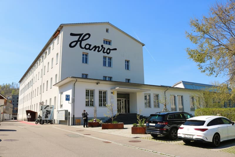 White building with name Hanro, large windows, entrance, person, 2 cars parked in front, trees, bushes, motorcycle