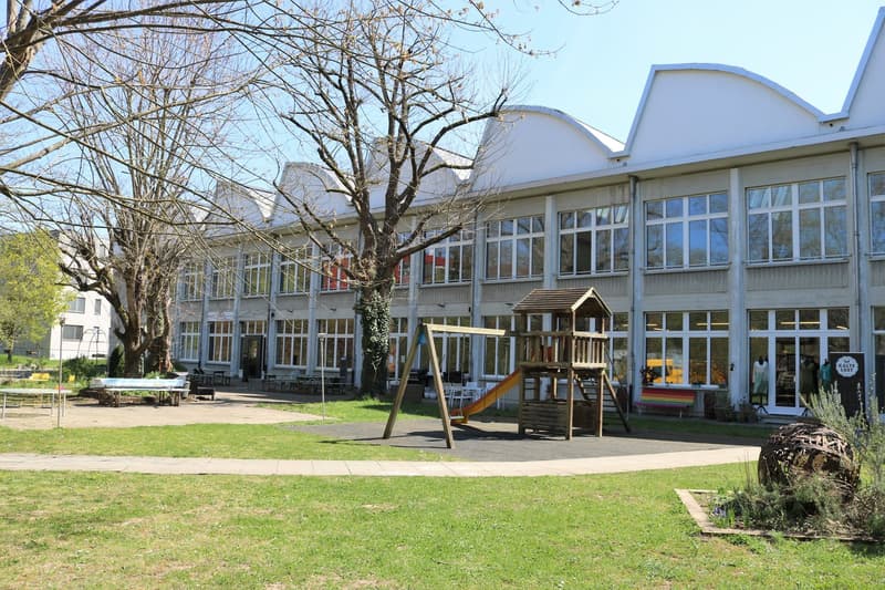 The building is white with arched roof sections. The building features many windows with white frames and glass panes. The entrance is wide with glass doors. There is a playground outside the building with a slide and a climbing frame. The ground is paved