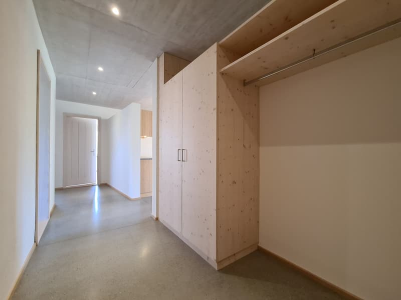 Empty hallway with white walls and gray floors, wooden cabinet on the right, small room visible at the end.