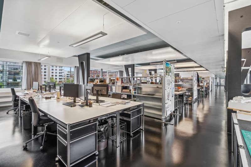 Large open-plan office space with modern furniture and equipment, including desks, computers, and shelving units. The space has high ceilings, large windows providing natural light, and a clean, minimalist design.