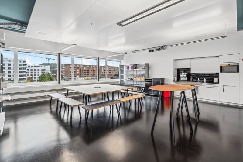 Large open office space with floor-to-ceiling windows, providing a view of the surrounding buildings. The room is equipped with modern furniture, including tables, chairs, and shelving units. The space appears to be designed for collaborative work or meet