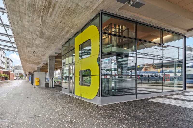 The image shows the exterior of a modern commercial building with a large yellow letter 'B' displayed prominently on the glass facade. The building has a concrete ceiling and a paved outdoor area in front. The glass walls allow for a view of the interior,