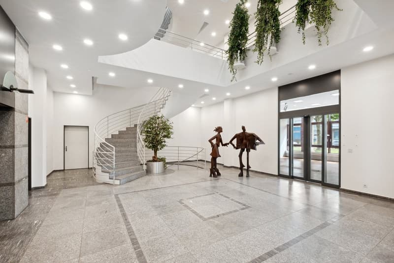 Spacious lobby with spiral staircase, potted plants, and abstract metal sculptures. The floor is made of gray granite tiles, and the ceiling has recessed lighting. Large glass doors lead to an outdoor area or balcony.
