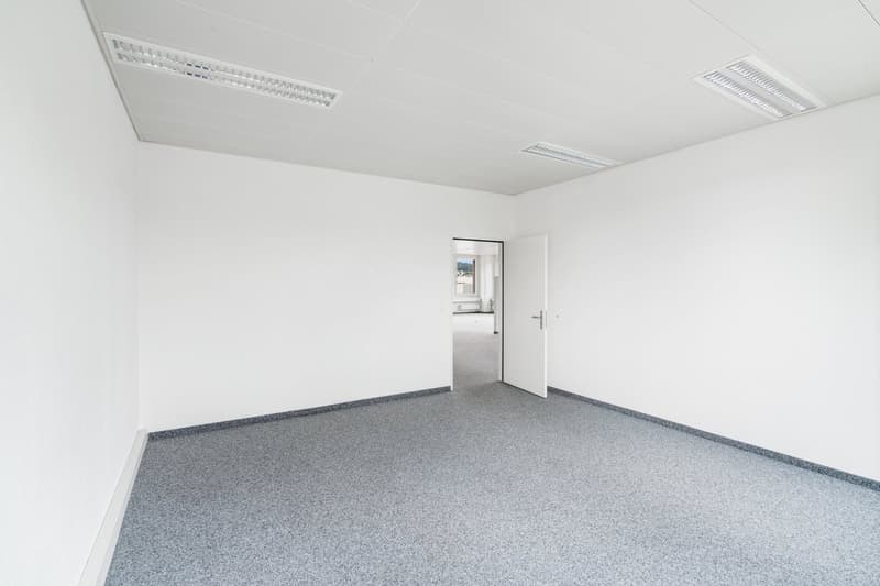 Empty office space with white walls, gray carpeted floor, and a door leading to another room. The ceiling has ventilation grilles.