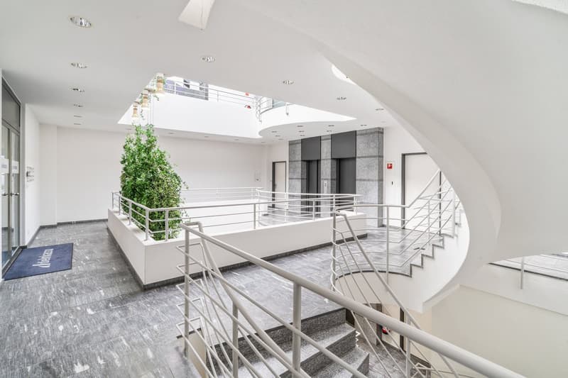 Spacious lobby area with high ceilings, white walls, and a balcony overlooking the lower level. The space features modern metal railings, stairs, and a decorative plant in the center.