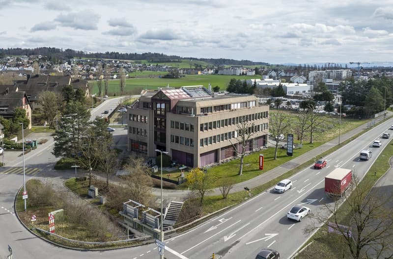 Office building, multiple floors, surrounded by trees and grass, located near a road with cars passing by, and a crane in the distance.