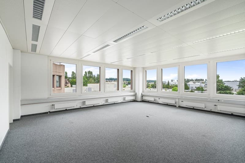 Large open office space with floor-to-ceiling windows providing a panoramic view of the surrounding landscape. The room has a neutral color scheme with white walls and gray carpeting. The ceiling features recessed lighting and ventilation systems.
