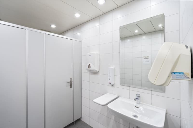Bathroom with white tiles, sink, mirror, soap dispenser, and toilet paper dispenser.