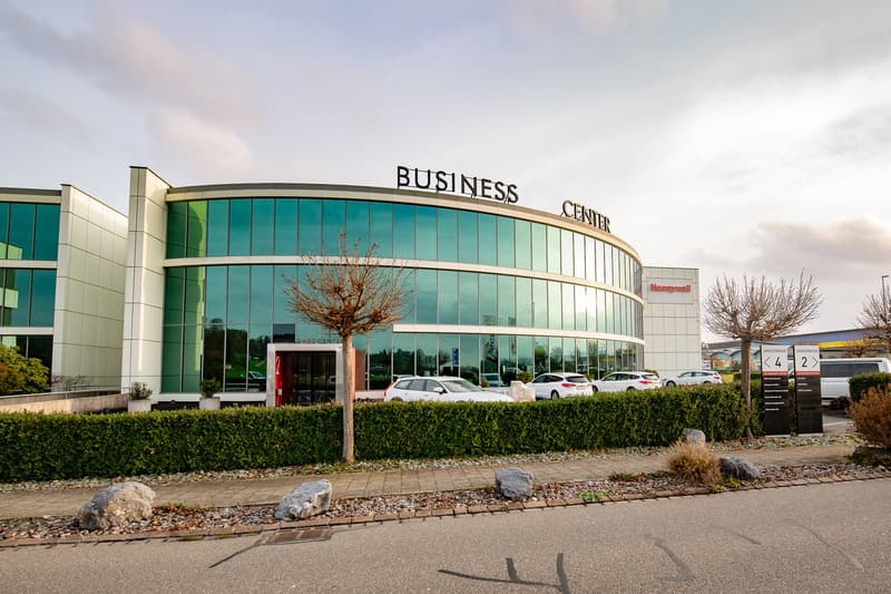 A large, multi-story glass and concrete building with the text 'BUSINESS CENTER' prominently displayed. The building has a curved facade and is surrounded by a parking lot with several cars parked. There is landscaping with trees and shrubs around the bui