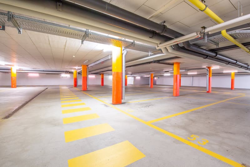 Large indoor parking garage with concrete floor, yellow and orange painted columns, and yellow striped markings on the floor indicating parking spaces.