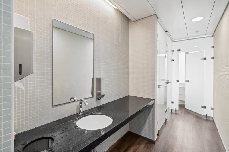 white tiled walls, black marble countertop, white sink, faucet, glass mirror, paper towel dispenser, soap dispenser, open shower area