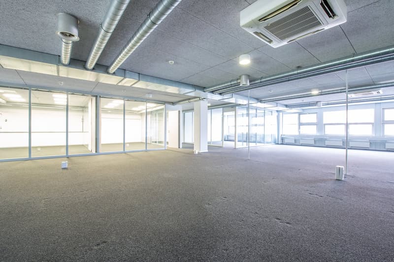 Large room with a glass wall and windows, carpeted floor, gray ceiling with metal pipes, ceiling-mounted air conditioners, lights, metal poles, and small white boxes.