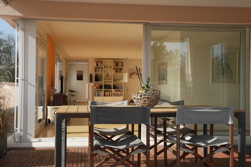 Open-concept living space with large windows, wooden dining table and chairs, bookshelves, and decorative accents