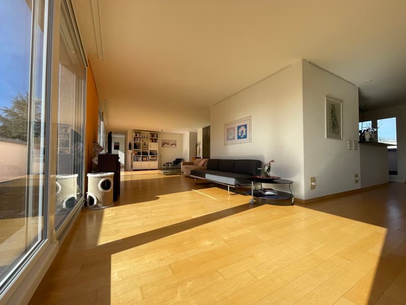 Spacious living room with large windows, hardwood floors, built-in shelving, and a sofa set. The room has an open layout and appears to have access to a balcony or terrace.
