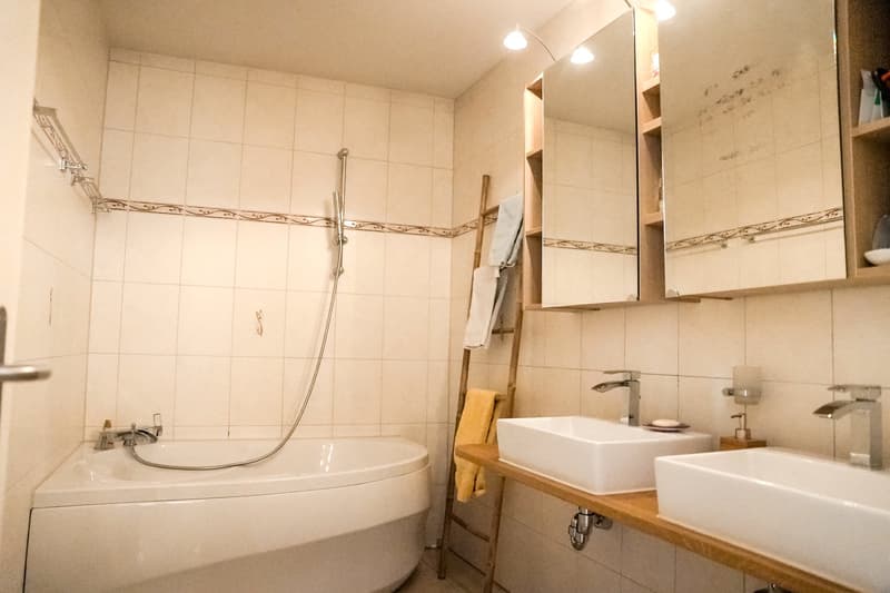 Tiled bathroom with a bathtub, vanity with two sinks, and a mirrored cabinet above. There is a towel rack and a washing machine visible in the room.