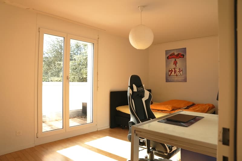Spacious bedroom with large windows, hardwood floor, gaming chair, desk, and a movie poster on the wall.