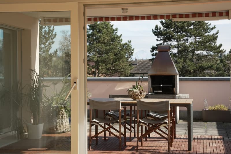 Covered patio with a fireplace, wooden dining table and chairs, potted plants, and a view of the surrounding pine trees through the large windows